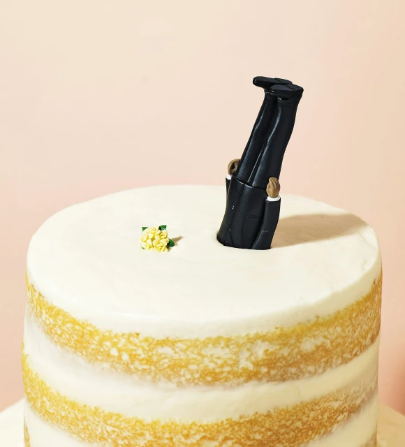 Novel wedding cake topper with groom figurine head-first in cake, small yellow flowers, against a plain pink backdrop, studio lit.