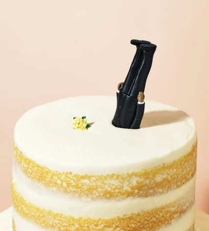Novel wedding cake topper with groom figurine head-first in cake, small yellow flowers, against a plain pink backdrop, studio lit.