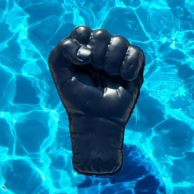 Inflatable black fist floats on the surface of a pool with clear blue water, creating rippling reflections and abstract patterns.