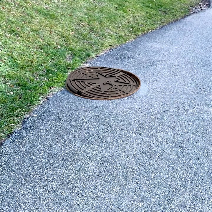 Brown manhole cover with a star design embedded in a concrete sidewalk, bordered by a grassy area in an outdoor setting.