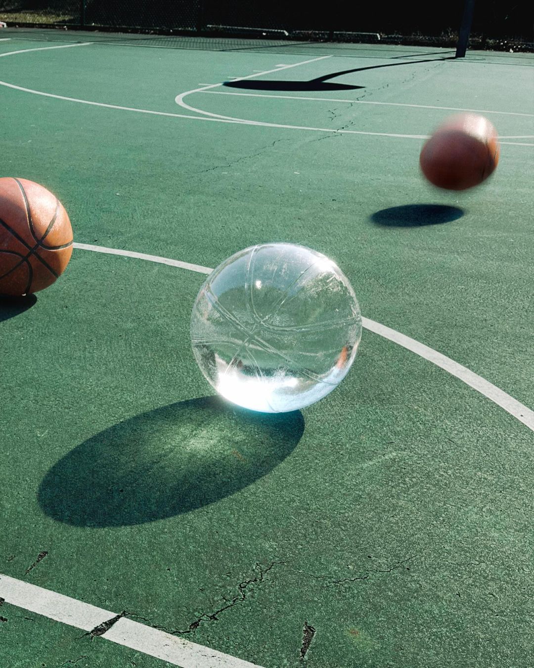 Basketball court with a clear crystal ball reflecting basketballs, one in motion, creating a surreal and dynamic composition outdoors.