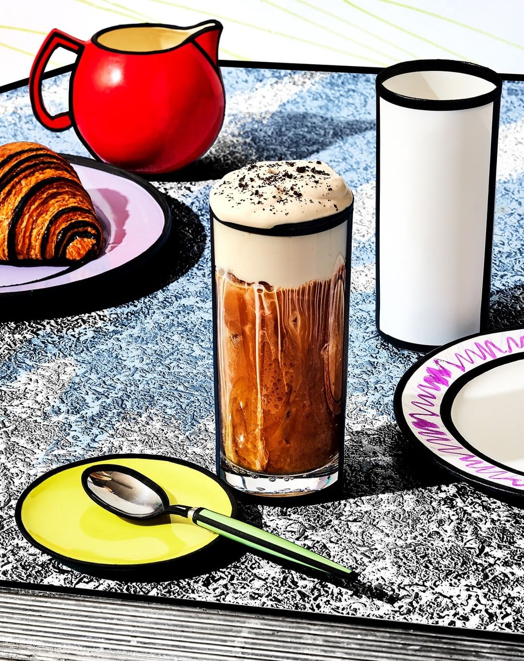 Stylized illustration of breakfast with croissant, chocolate iced coffee, spoon, red pitcher, and white cup, cartoonish, colorful.