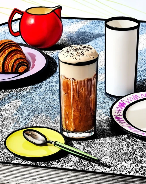 Stylized illustration of breakfast with croissant, chocolate iced coffee, spoon, red pitcher, and white cup, cartoonish, colorful.