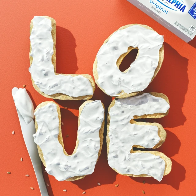 Overhead shot of sugar cookies shaped into 'LOVE' with white frosting and a knife on a red background with a cream cheese package.