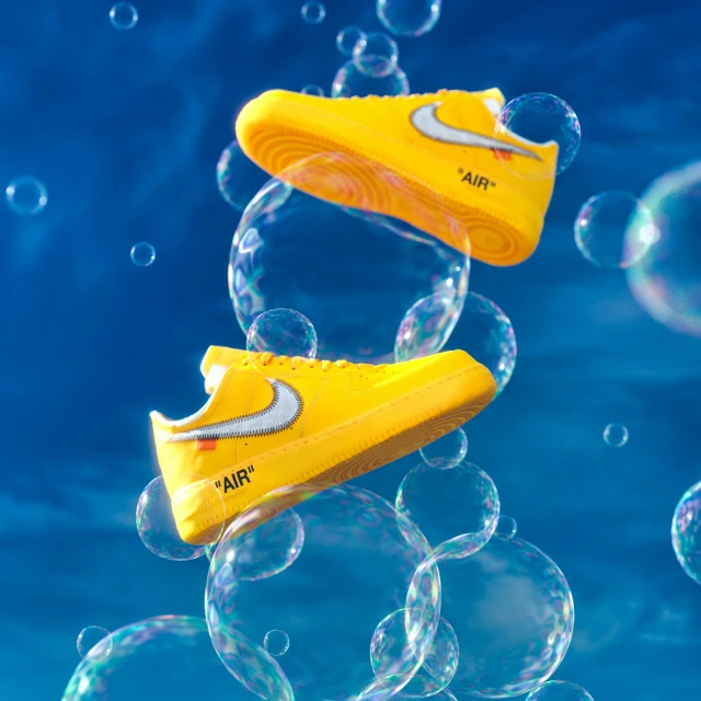 Two yellow Air Force 1 sneakers float among transparent soap bubbles against a vibrant blue sky background in a product shot.