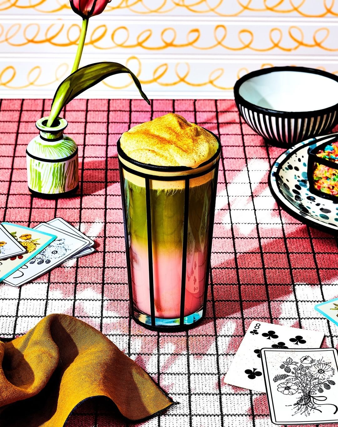 Artistic still life photo of a colorful layered drink with foam in a glass, arranged with playing cards, flowers, and striped dishware.