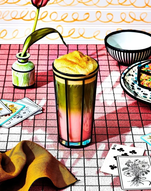 Artistic still life photo of a colorful layered drink with foam in a glass, arranged with playing cards, flowers, and striped dishware.
