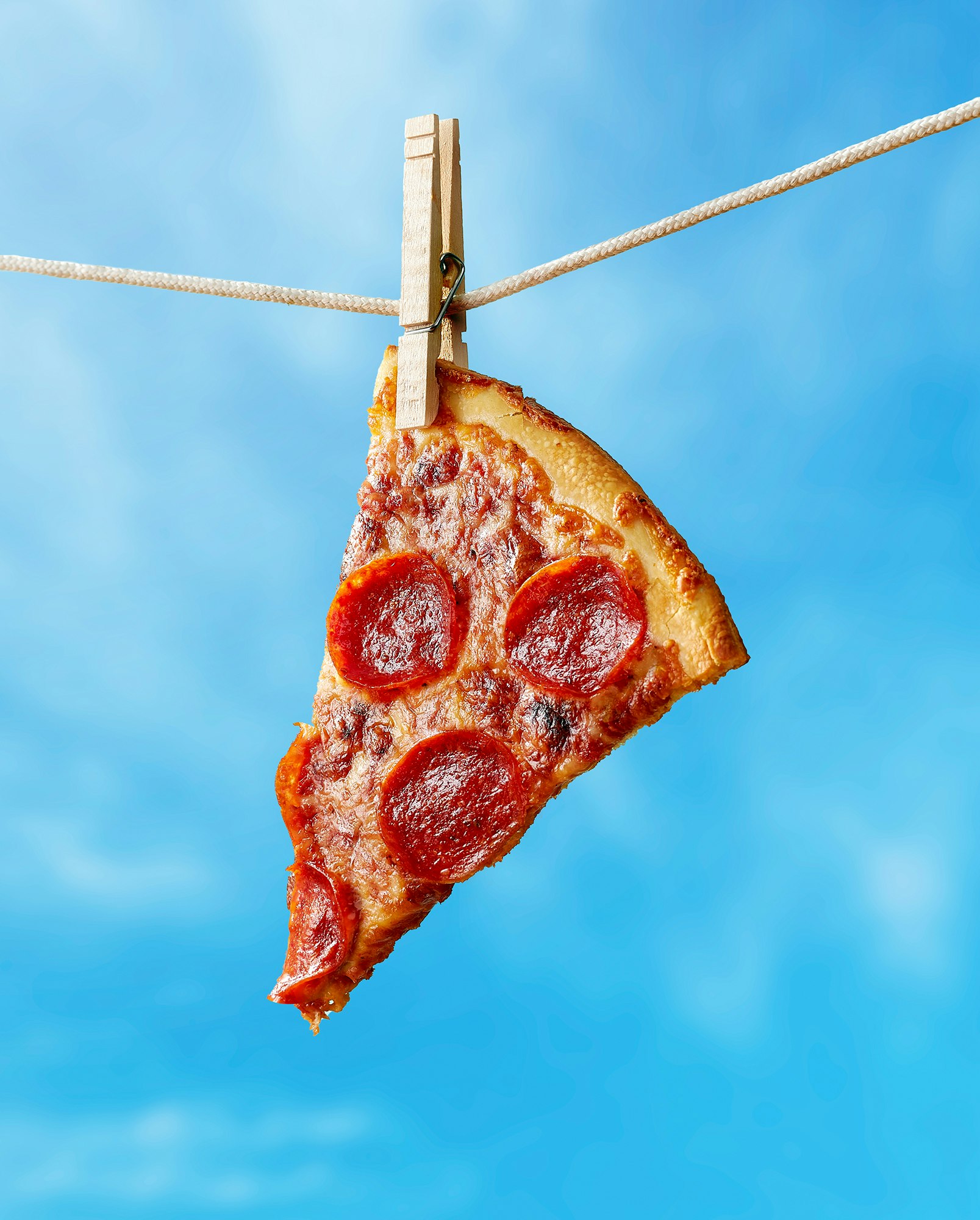 Slice of pepperoni pizza hanging from a clothesline with a clothespin against a vibrant blue sky. Food photography, high angle.
