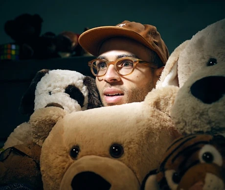 Portrait of an adult male wearing glasses and a baseball cap, surrounded by tan and white stuffed animals indoors in dim lighting.