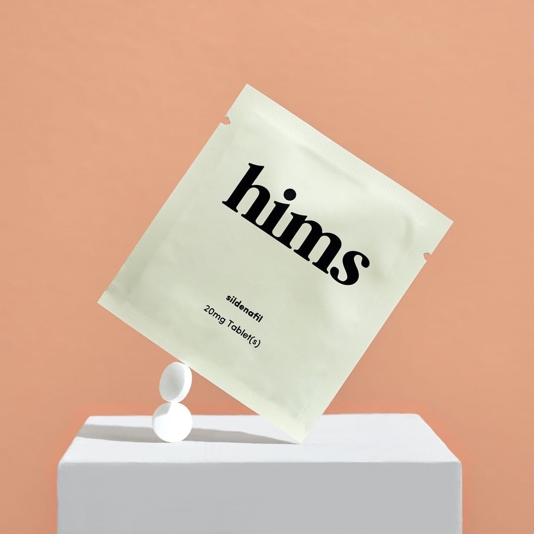 Still life studio shot of a packet of sildenafil pills balanced on two white pills sitting on a white cube against a peach background.