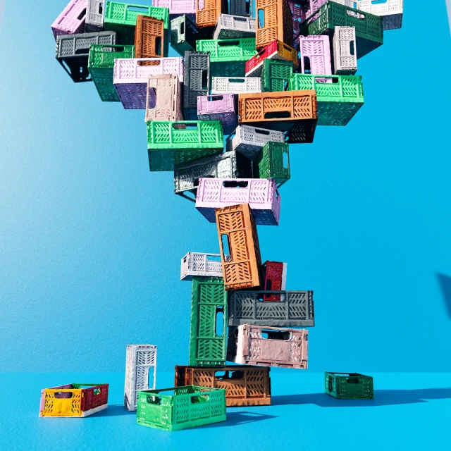 Stack of colorful plastic crates balanced on a blue background, creating a whimsical and playful visual composition.