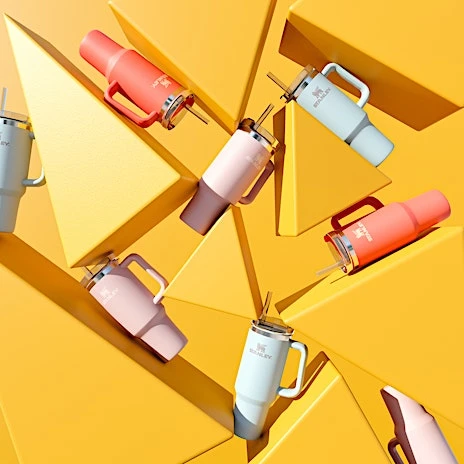 Several Stanley mugs in varying colors are arranged against a bright yellow geometric background, creating a visually striking product display.