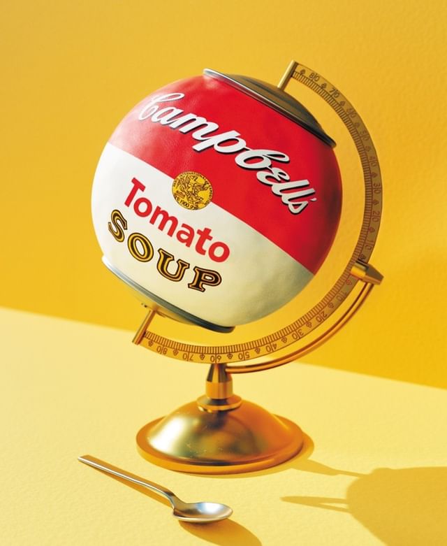 A Campbell's Tomato Soup can is creatively displayed as a globe, supported by a gold stand, accompanied by a spoon on a yellow background.