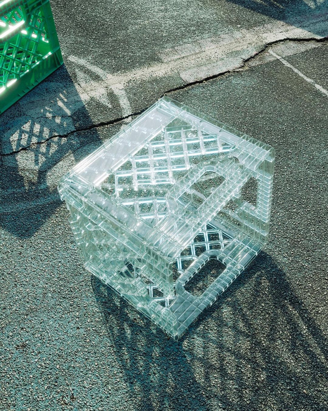 Clear plastic milk crate sitting on cracked asphalt, casting shadows. One green milk crate visible in the upper left corner.