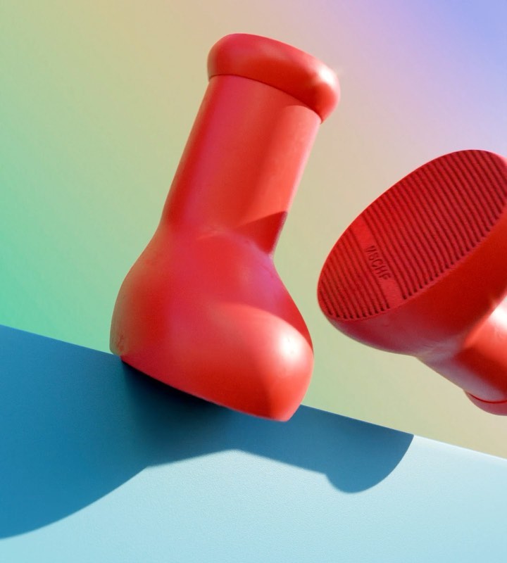 Two bright red rubber boots displayed on a blue surface against a pastel gradient background in a studio product shot.