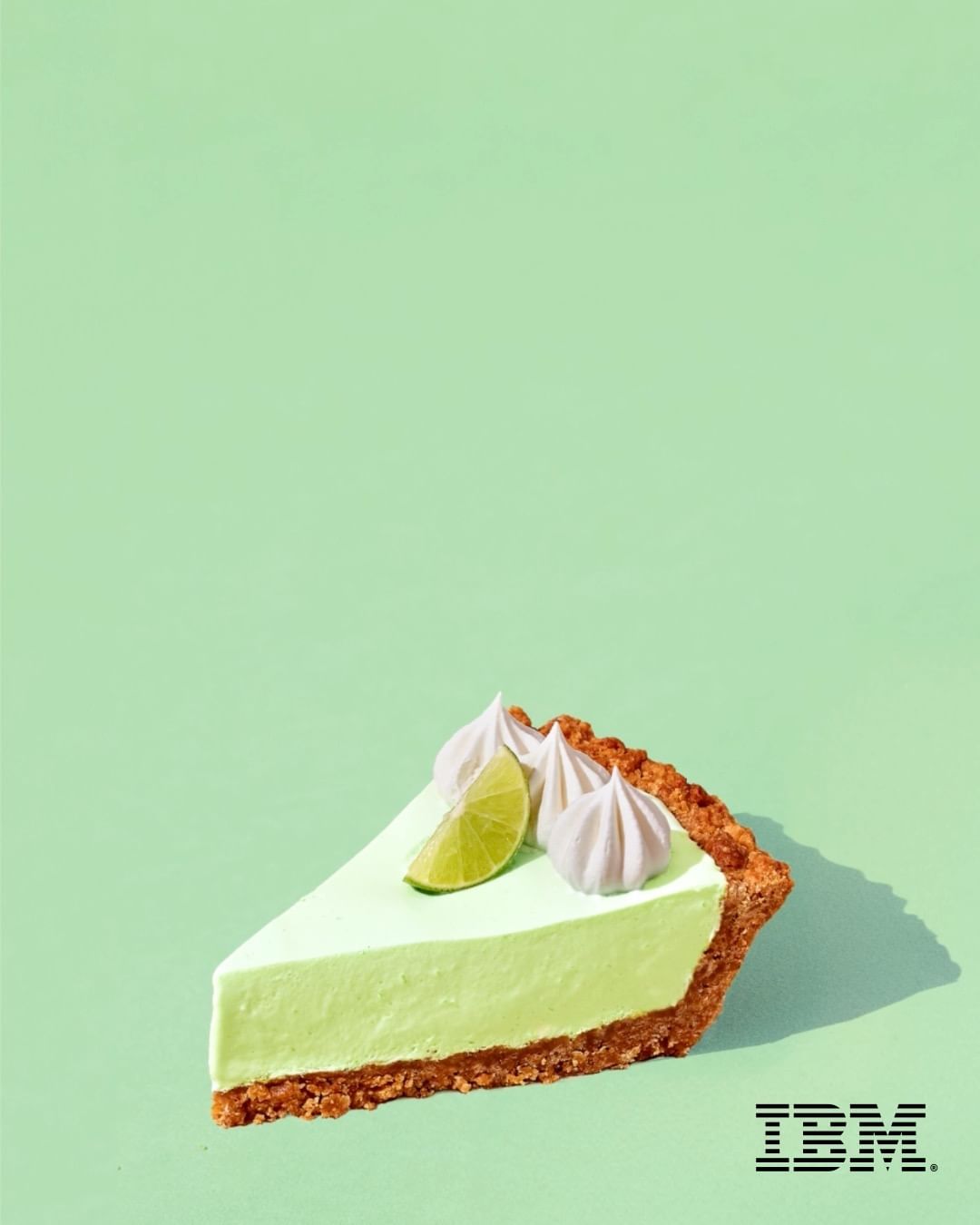 A slice of key lime pie on a light green background, topped with lime and meringue, sitting in a studio environment with visible branding.