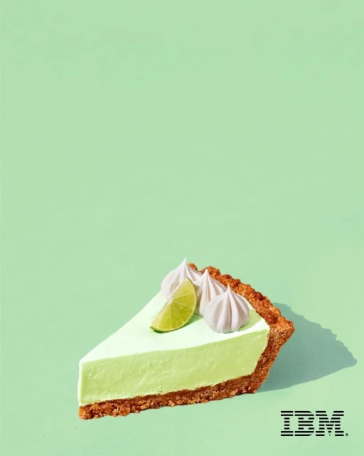 A slice of key lime pie on a light green background, topped with lime and meringue, sitting in a studio environment with visible branding.