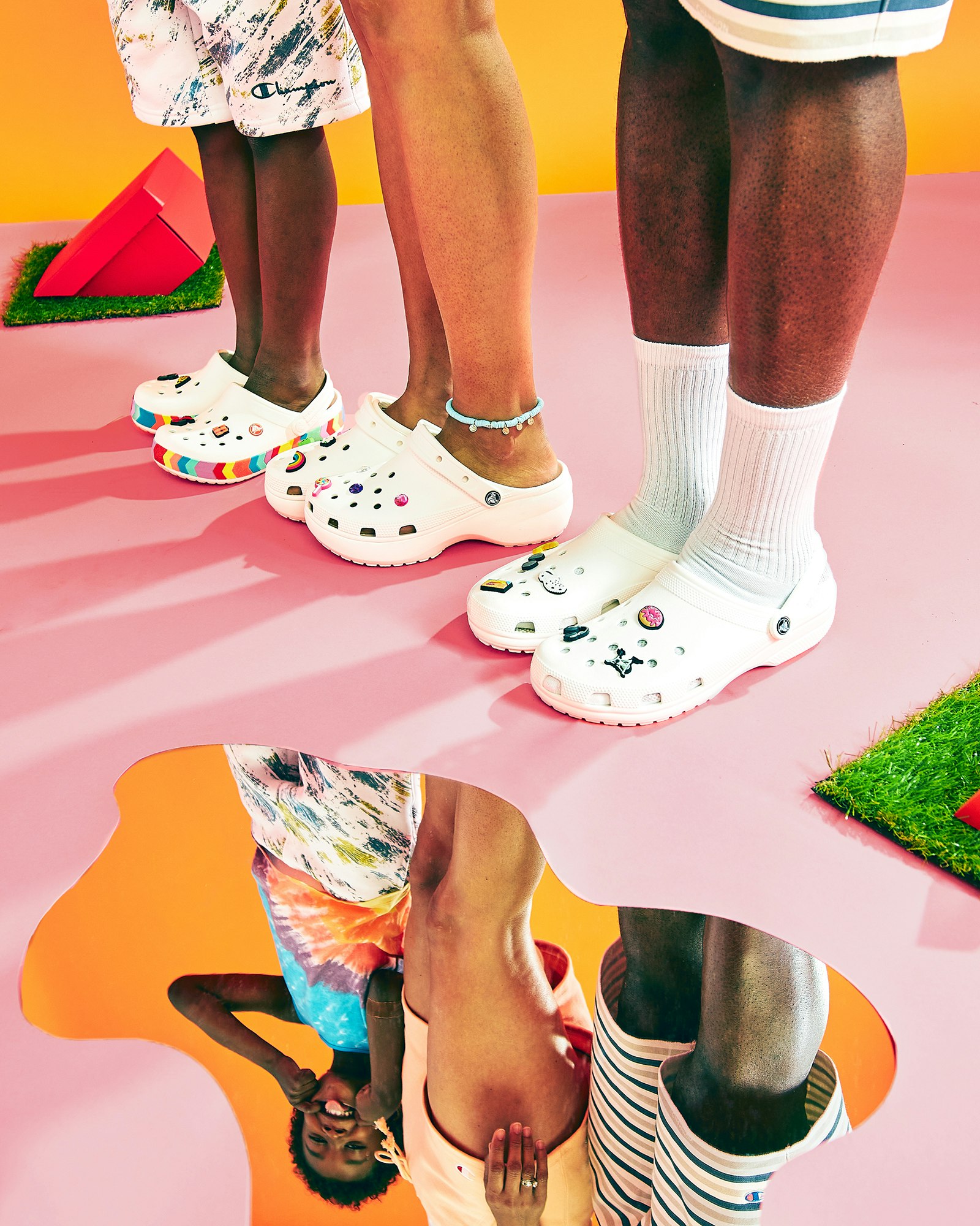 Three people wearing white Crocs stand in a studio with pink and orange background, reflected in a puddle-like mirror on the floor.
