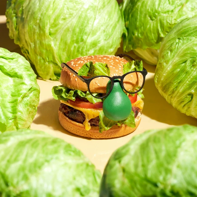 Hamburger 'disguised' with glasses and a fake nose surrounded by iceberg lettuce heads on a tan background in a studio setting.