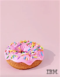 Overhead shot of a pink frosted doughnut with colorful sprinkles on a light pink background with shadow and the IBM logo at the bottom.