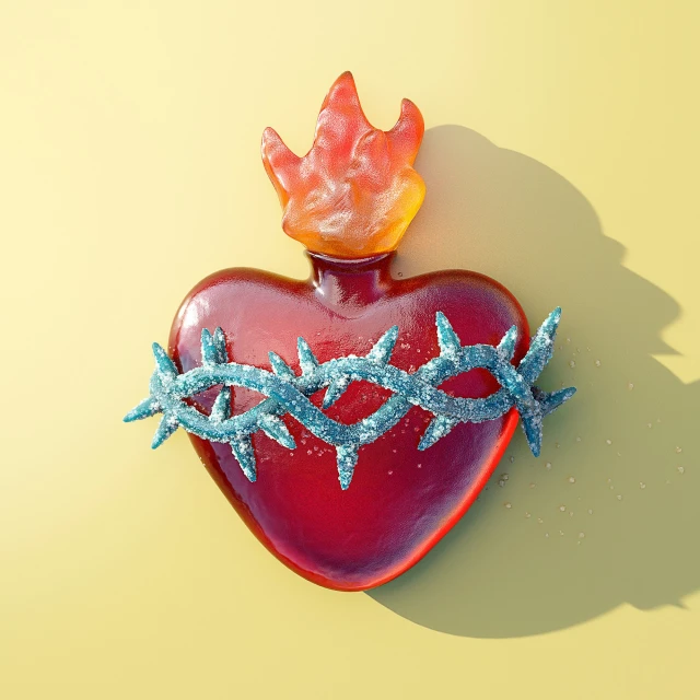 Red heart art piece with a crown of thorns and flame atop, casting a shadow on a pale yellow background, art photography.