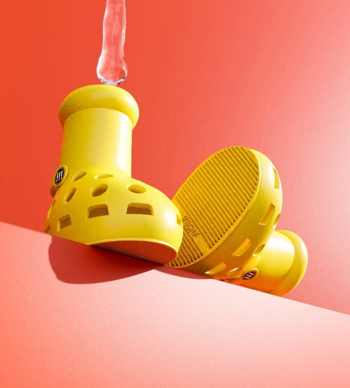 Elevated studio shot of a pair of yellow rubber boot-shaped sandals, one upright with water pouring into it against orange background.