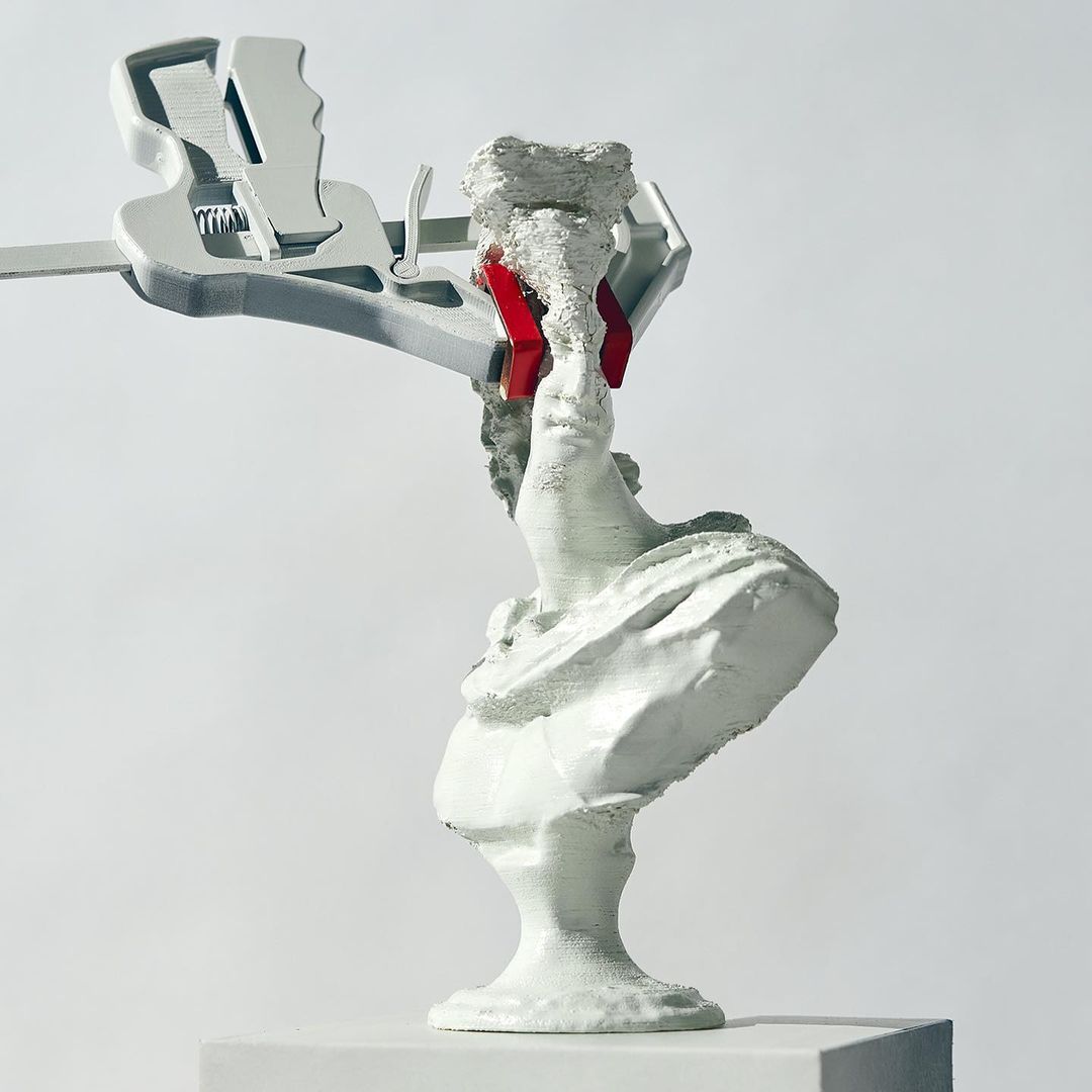 Bust sculpture in white paint, held by a metal wrench with red accents, displayed on a white plinth against a white backdrop.