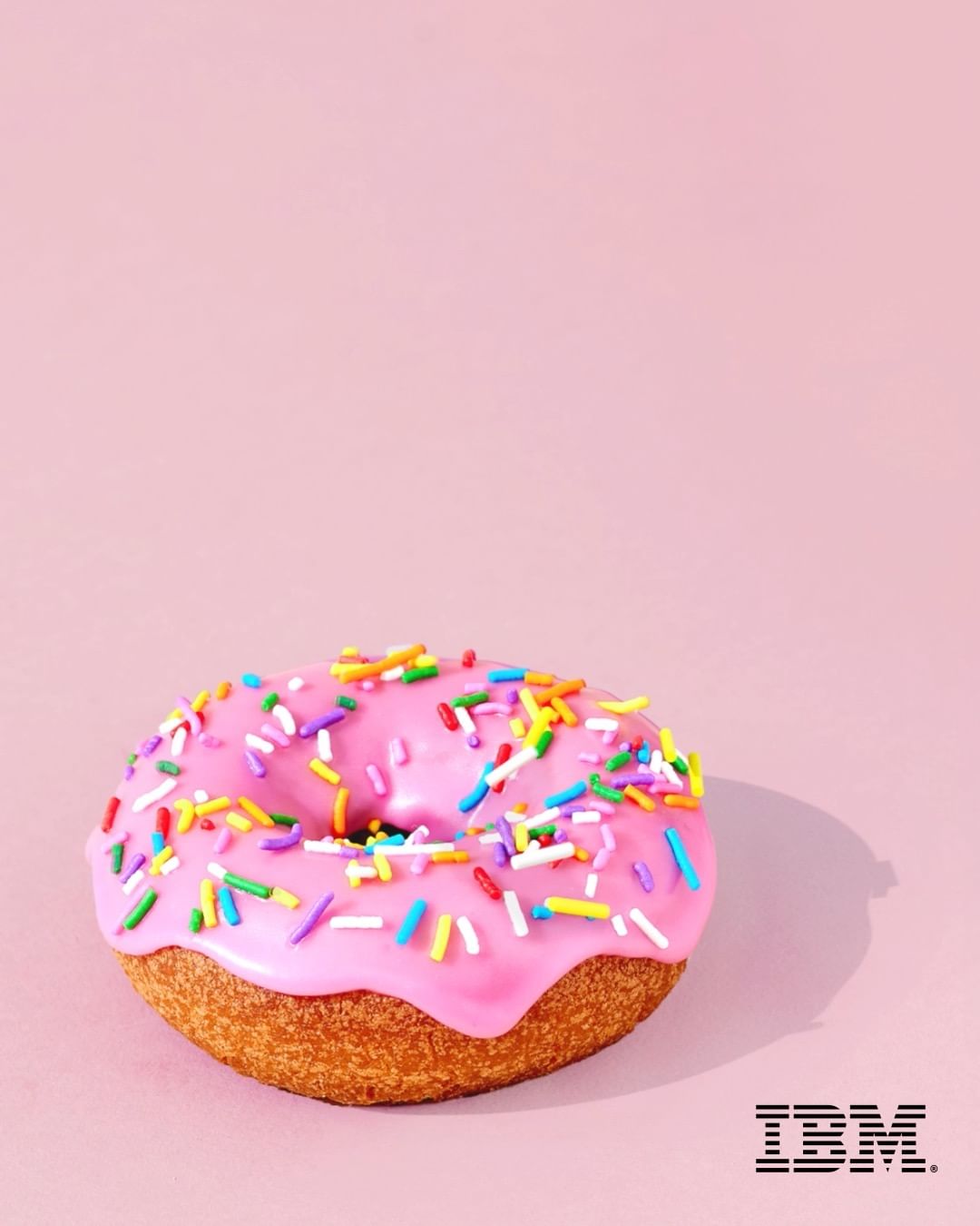 Eye-level studio shot of a pink donut with colorful sprinkles on a light pink backdrop with the IBM logo in the bottom right corner.