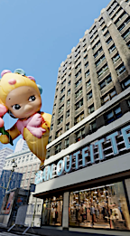 Macy's Thanksgiving Day Parade featuring a large character balloon floating near a tall building with signage on a sunny day.