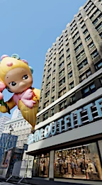 Macy's Thanksgiving Day Parade featuring a large character balloon floating near a tall building with signage on a sunny day.