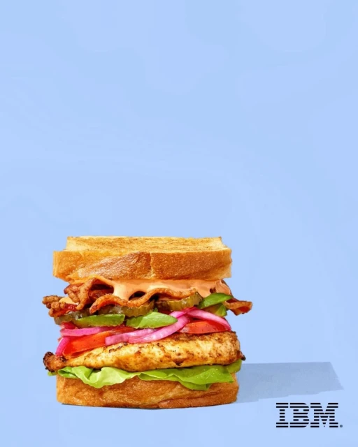 Stacked chicken sandwich with toast, bacon, tomato, onion, pickles and lettuce against a light blue backdrop, food photography.