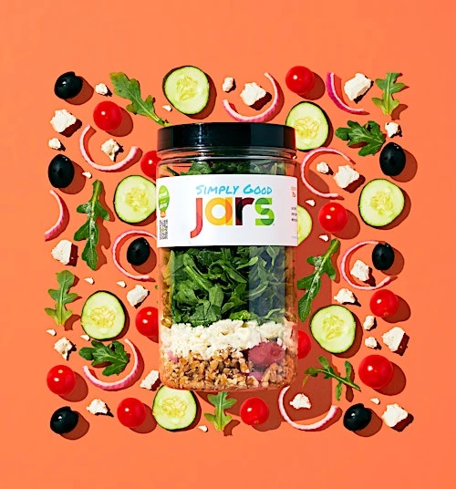 Overhead shot of a 'Simply Good Jars' salad jar surrounded by fresh ingredients: tomatoes, cucumbers, olives, onions, and greens, on orange.
