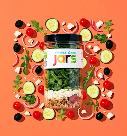 Overhead shot of a 'Simply Good Jars' salad jar surrounded by fresh ingredients: tomatoes, cucumbers, olives, onions, and greens, on orange.