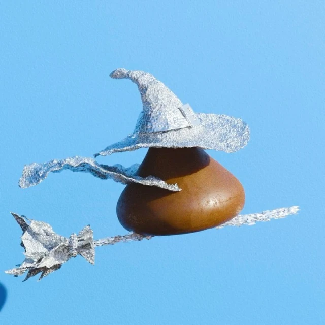 A chocolate candy kiss wearing a silver glittery witch hat and riding a silver glittery broom on a light blue background. Halloween themed.