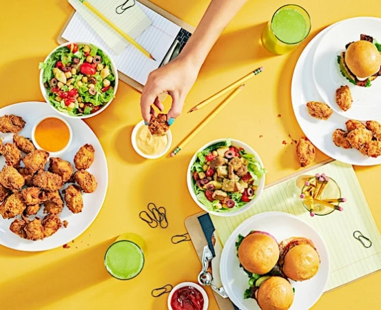 Overhead shot of hands reaching for food, with plates of fried foods, salads, smoothies, and stationery on a yellow surface.