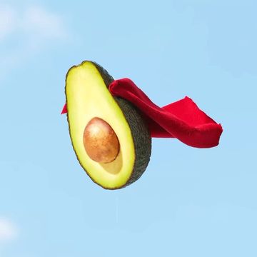 Avocado half with pit and red cape flying against a light blue sky background, vibrant food photography for advertising.