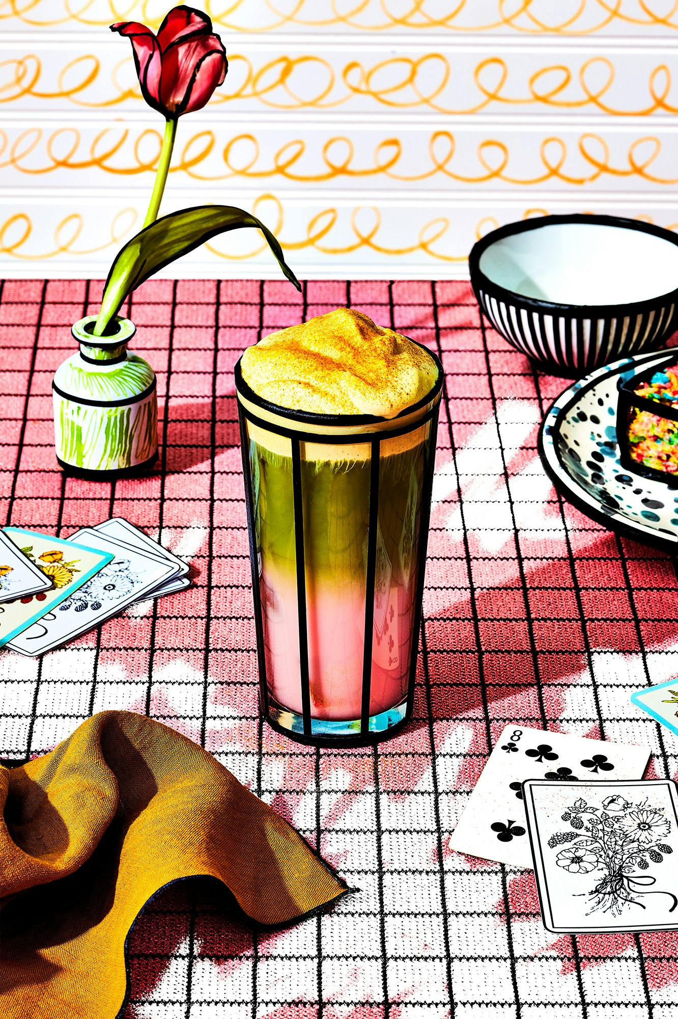 Whimsical still life: a stylized flower in a vase and a mixed drink in a patterned glass on a red and white checkered tablecloth.