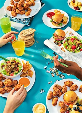 Overhead food spread featuring fried chicken, burgers, salads, and drinks on a bright blue surface by Andre Rucker