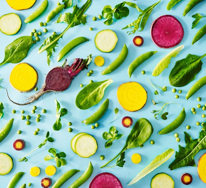 Overhead shot of a flat lay food arrangement, showcasing fresh green vegetables such as peas, zucchinis, beetroots, and lettuce on a light blue surface.