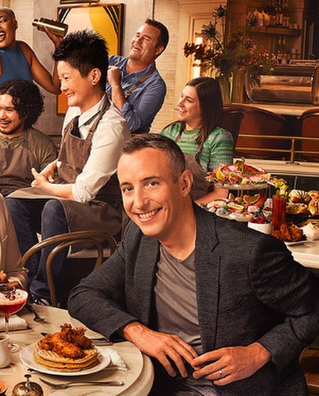 Group of people seated at a table with food and drinks, featuring a man in a suit jacket smiling directly at the camera.