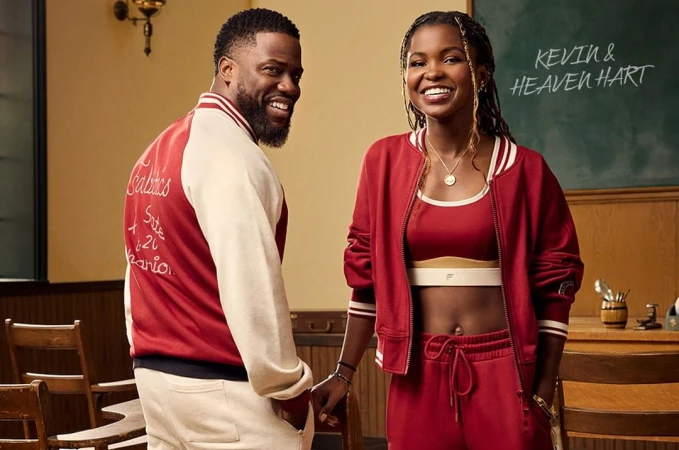 Kevin and Heaven Hart pose in a classroom setting wearing matching red outfits. Kevin wears a letterman jacket; Heaven wears a track suit.