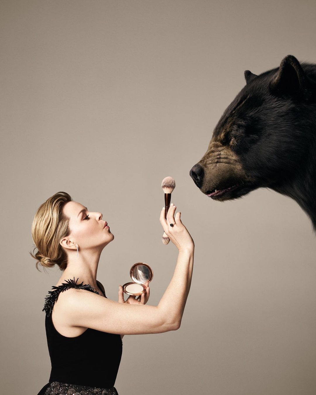 Elegant woman in black dress holds makeup brush and compact near a large black bear head in a studio setting on a gray background.