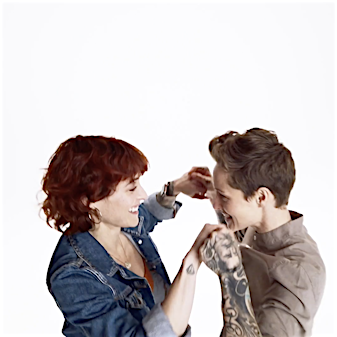 Two adult women laugh together, holding hands in a bright photo studio. One woman has auburn curly hair; the other has short brown hair and tattoos.