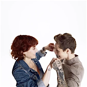 Two adult women laugh together, holding hands in a bright photo studio. One woman has auburn curly hair; the other has short brown hair and tattoos.