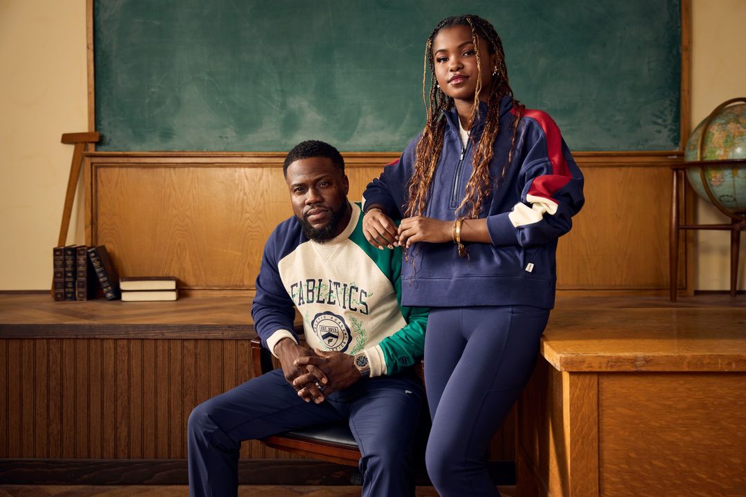 Kevin Hart and a young woman are posing together in front of a school desk. The pair are wearing Fabletics athletic clothing.