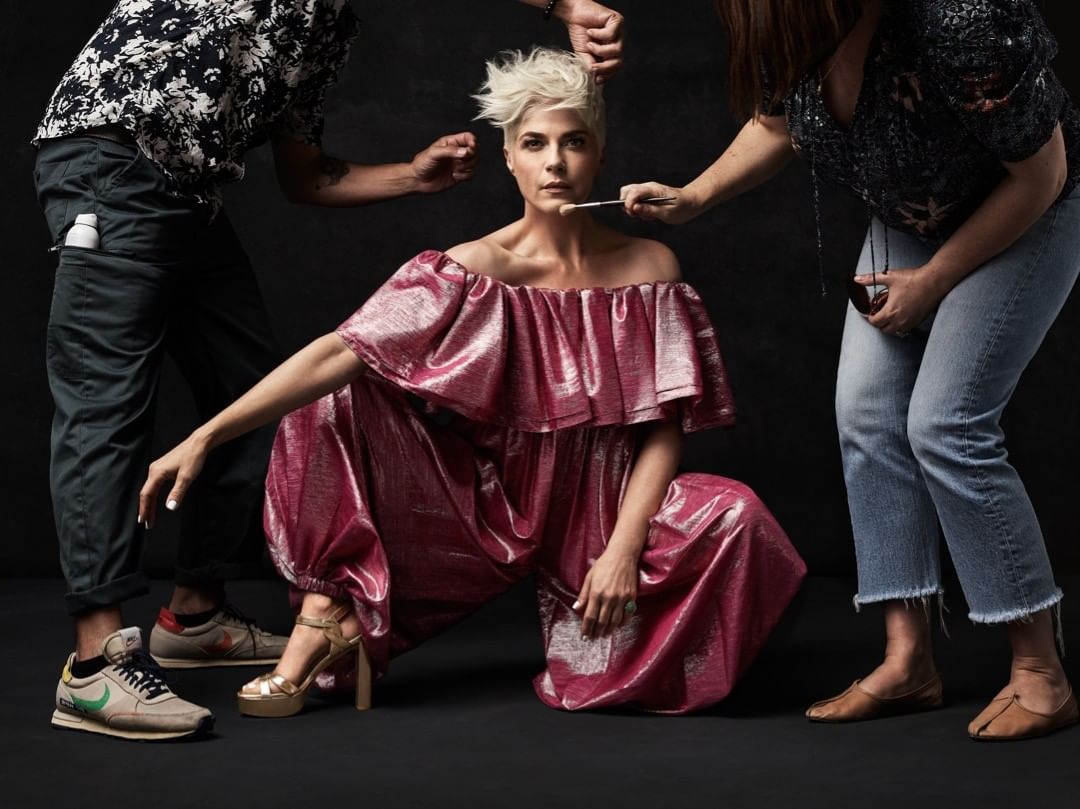 Actress Selma Blair in a pink metallic jumpsuit, kneeling in a studio with two other people attending to her hair and makeup.