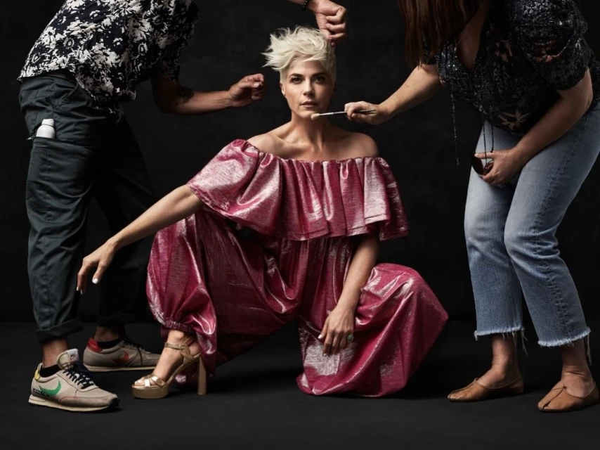 Actress Selma Blair in a pink metallic jumpsuit, kneeling in a studio with two other people attending to her hair and makeup.