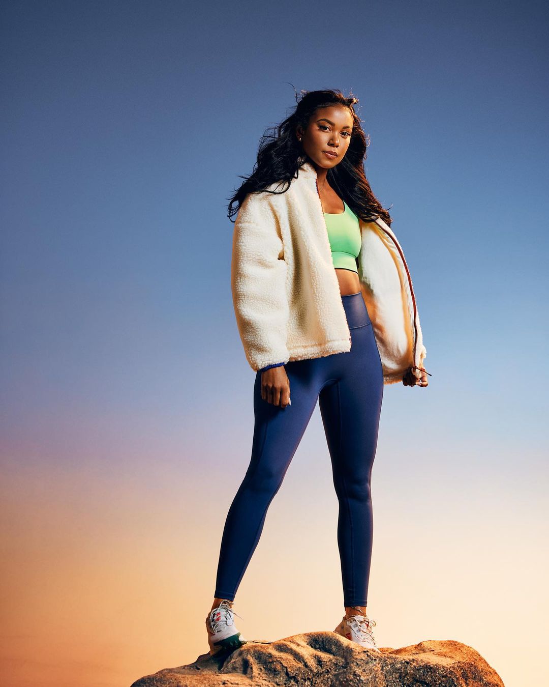 A confident woman in athletic wear strikes a pose on a rock against a blue and orange sky, wearing leggings, sneakers, and a fleece jacket.