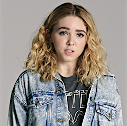 Young woman with curly blonde hair in a denim jacket and a Led Zeppelin t-shirt poses against a light gray background in a studio portrait.