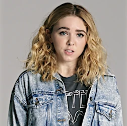 Young woman with curly blonde hair in a denim jacket and a Led Zeppelin t-shirt poses against a light gray background in a studio portrait.
